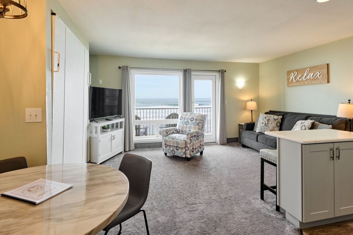Living room with ocean-view balcony: gray sofa, patterned chair, media stand, and large windows showing the sea.