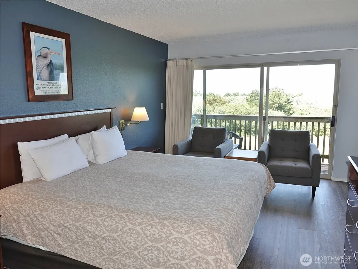 Hotel room with a large bed, blue accent wall, framed bird poster, and a balcony with two gray chairs overlooking trees.