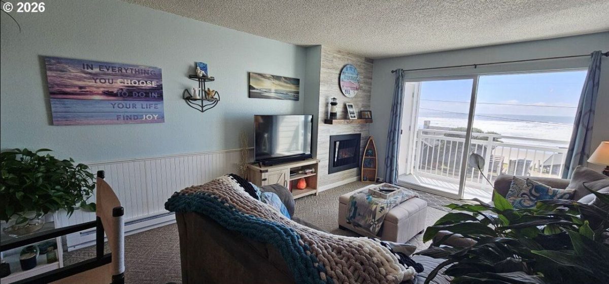 Cozy living room with a knit throw on the sofa, TV stand, and a sliding glass door to a balcony overlooking the beach.